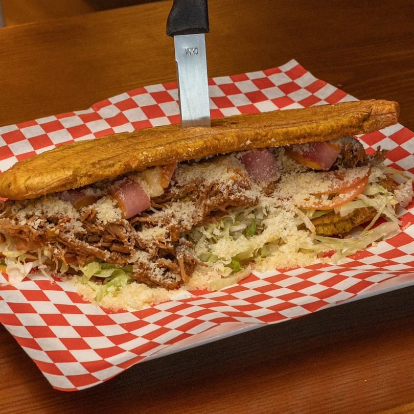 A Colombian patacón sandwich with shredded meat, lettuce, tomato, cheese, and a knife on checkered paper.
