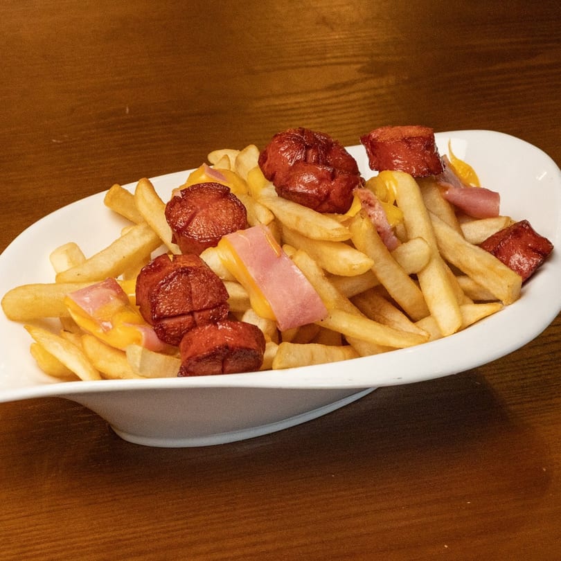 A white plate of golden french fries topped with sliced hot dogs, ham, and melted cheese.